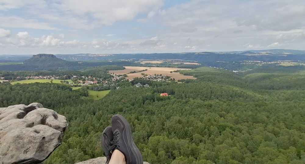 Das Bild zeigt eine atemberaubende Aussicht von einem felsigen Aussichtspunkt auf eine weitläufige Landschaft mit einem Dorf, Wäldern und einem Tafelberg im Hintergrund.

Das Bild zeigt die Füße einer Person, die auf einem felsigen Vorsprung sitzt und die Aussicht genießt. Die Person trägt schwarze Laufschuhe. Der Vordergrund besteht aus einem großen, hellgrauen Felsen mit unregelmäßiger Form und mehreren Löchern und Vertiefungen. Im mittleren Grund und Hintergrund erstreckt sich ein grünes Waldgebiet, das sich bis zum Horizont erstreckt.  Man kann einzelne Bäume und Lichtungen erkennen. In der mittleren Entfernung ist ein kleines Dorf mit Häusern verschiedener Größen und Farben zu sehen.  Felder, wahrscheinlich Ackerland, sind zwischen dem Wald und dem Dorf eingezwängt. Im Hintergrund ist ein markanter, flacher Tafelberg zu erkennen, der sich deutlich von der umliegenden Landschaft abhebt. Der Himmel ist leicht bewölkt, mit einem Mix aus weißen und hellgrauen Wolken, was zum Gesamtbild einer ruhigen und friedlichen Atmosphäre beiträgt.  Die Stimmung des Bildes ist entspannt, friedlich und vermittelt ein Gefühl von Freiheit und Weite. Die Farben sind überwiegend grün und grau, mit einem Hauch von Braun aus den Feldern und den rötlichen Dächern der Häuser.
