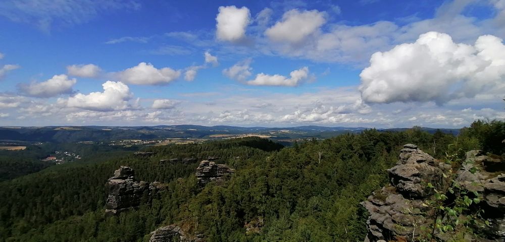 Eine weite Aussicht auf eine Landschaft mit Hügeln, Wäldern und Felsklippen unter einem blauen Himmel mit weißen Wolken.

Die Komposition der Szene zeigt eine Panoramaaussicht auf eine weitläufige Landschaft.  Die Perspektive ist von oben nach unten gerichtet, was eine umfassende Sicht auf die Region ermöglicht. Die verschiedenen Höhenstufen, die aus Wäldern, Tälern und Felsklippen bestehen, sind deutlich zu erkennen. Der Vordergrund wird von einer Reihe von felsigen Klippen und Bäumen eingenommen, die sich nach hinten in dichte Wälder ausdehnen.  Das mittlere und hintere Gelände ist von einer Reihe von Hügeln und Tälern geprägt, die sich ausdehnen und sich nach hinten in eine flachere Landschaft verwandeln.  Die weiten Blickwinkel vermitteln eine tiefe Perspektive.

Die wichtigsten Elemente sind die vielfältige Landschaft, einschließlich der Wälder, die sich in unterschiedlichen Grüntönen über die Szene erstrecken. Die Felsformationen im Vordergrund sind in dunklen Grautönen gehalten und setzen einen starken Kontrast zur üppigen Vegetation.  Die Größe und der Umfang der Landschaft sind ein auffälliges Merkmal, das eine bemerkenswerte räumliche Ausdehnung vermittelt. Die verschiedenen Farbwerte spiegeln die unterschiedlichen terrestrischen Umgebungen wider.

Die Landschaft präsentiert einen sonnigen Tag mit einem klaren Himmel und weißen Wolken. Die Beleuchtung ist gleichmäßig verteilt und beleuchtet die verschiedenen Merkmale der Landschaft detailreich und lebendig.  Die allgemeine Stimmung ist friedlich und ergreifend, was durch den Panoramablick und die natürliche Schönheit der Landschaft unterstrichen wird. 