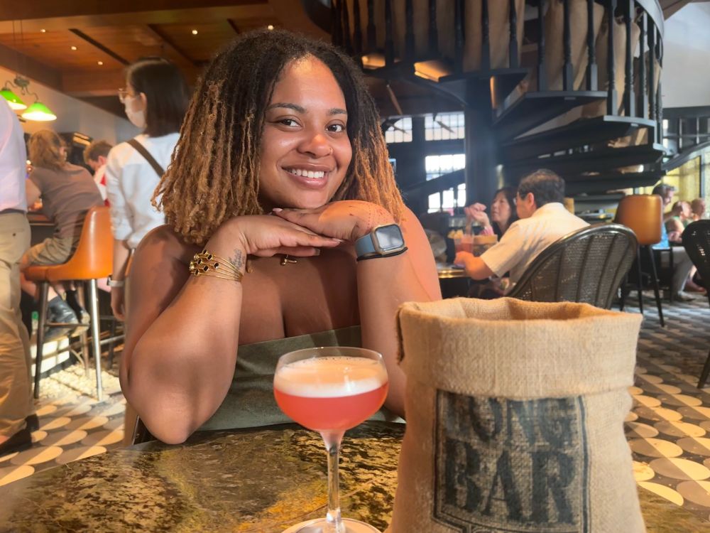 I’m at the Long Bar inside the Raffles Hotel in Singapore. I have a petite pink drink called a Sakura Sling. It was delicious. There is a bag of peanuts on the table that says Long Bar. And I’m sitting behind it lol