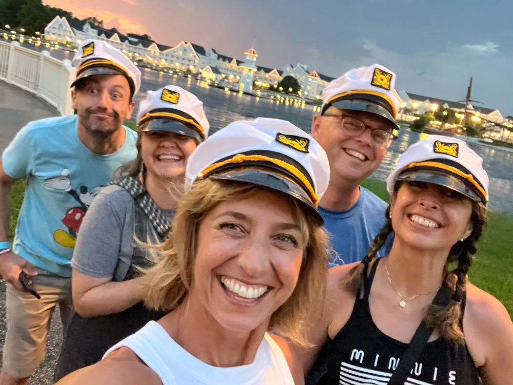 five friends smiling and having fun wearing silly yacht captain hats with Disney’s Boardwalk resort in the background