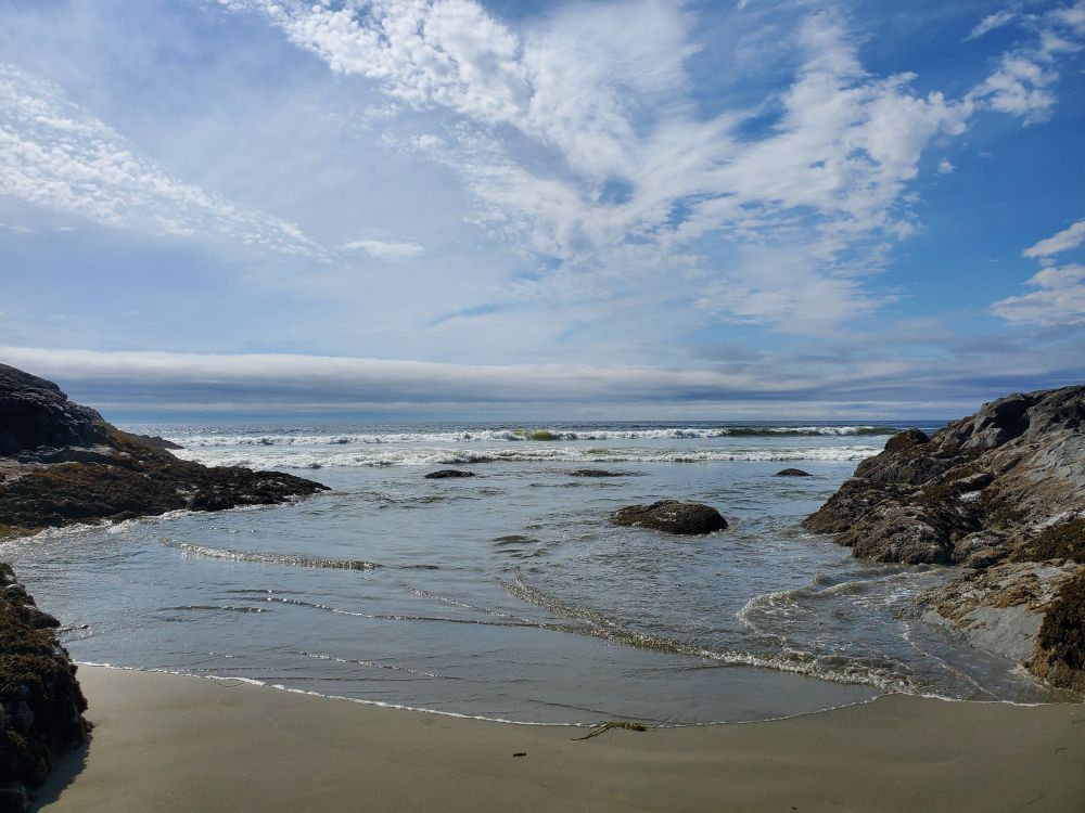 A shot of a beach in the sun. In the foreground, waves curl onto a sandy beach framed by dark, jagged rocks. In the background, the ocean meets windswept clouds streaking across a bright blue sky.