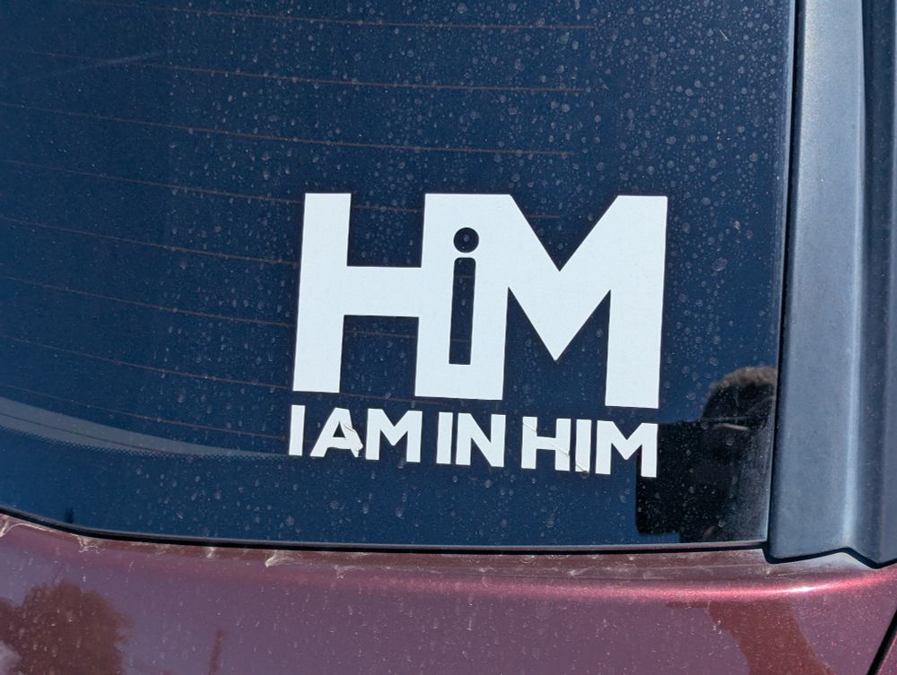 A decal on a car. It says "HiM" in the upper half, and "I AM IN HIM" in the lower half.