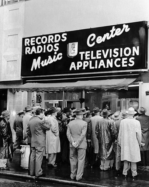 Black and white photo of a crowd completely filling the sidewalk in front of a storefront window, with a sign "Music Center, Records Radios Television Appliances"
