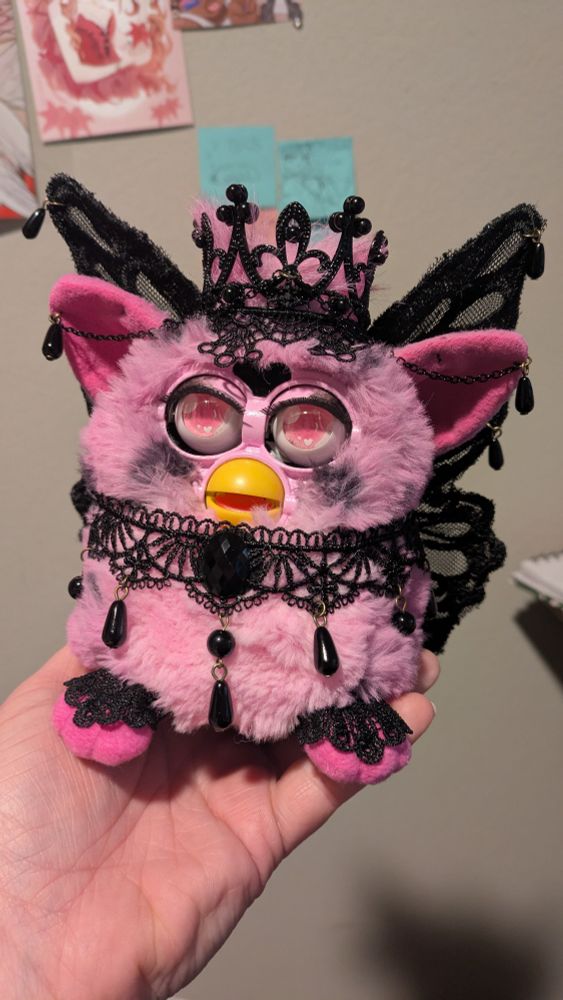 A custom of a furby; its pink with black spots and accessories, has black lace fair wings and a black crown