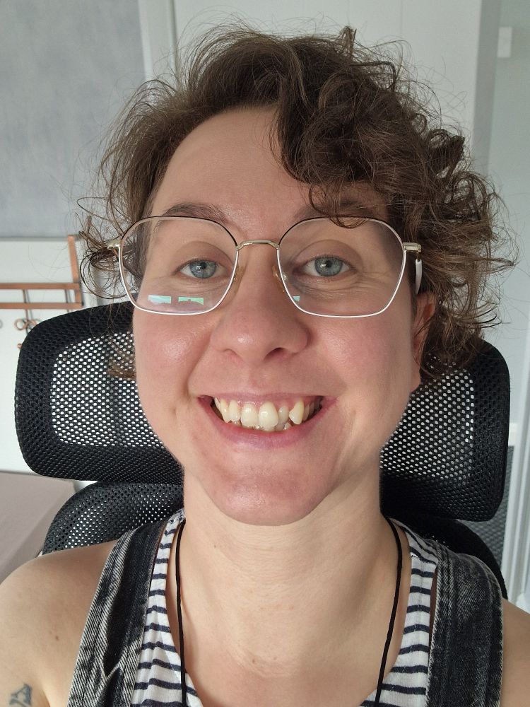 Selfie of Kat. He's wearing his usual black and white wire-rimmed glasses and is smiling at the camera, head resting on the head rest of a black mesh desk chair. He's wearing a white and black striped vest top and dark wash denim dungarees