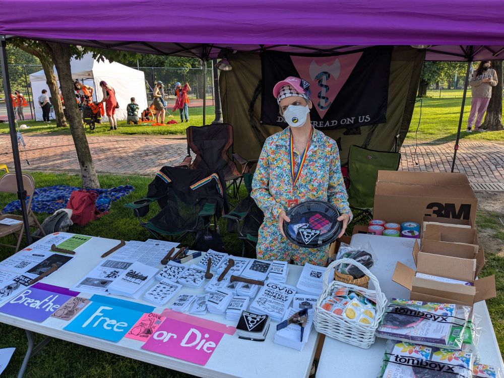 a woman of white wearing a pink hat, keffiyeh bandana, rainbow lanyard, and floral jumpsuit stands behind a table holding a platter of kn95 masks. on the tables there are various zines, condoms, masks, covid tests, and signage displaying the words "breathe free or die".