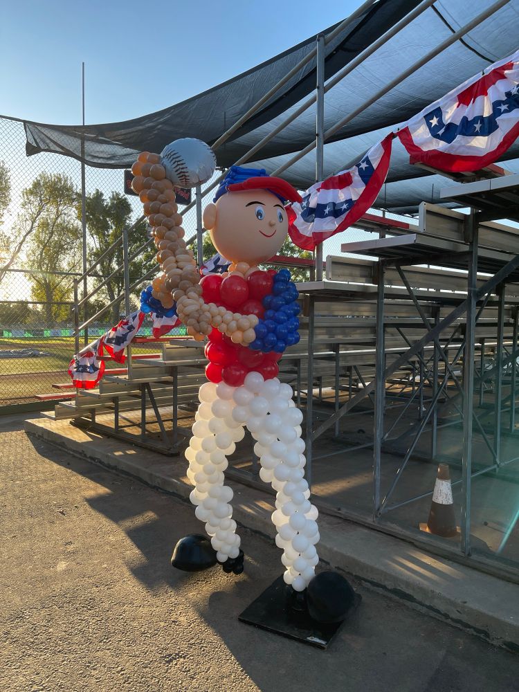 large balloon sculpture of a Little League player standing by bleachers nearby a baseball field. it wears a blue and red jersey with white pants 