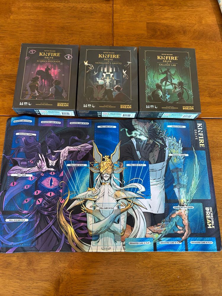 Three cars game boxes placed above a play mat depicting its main antagonists. The boxes are games in the Kinfire Delve Series: Scorn’s Stockade, Vainglory’s Grotto, and Callous’ Lab.