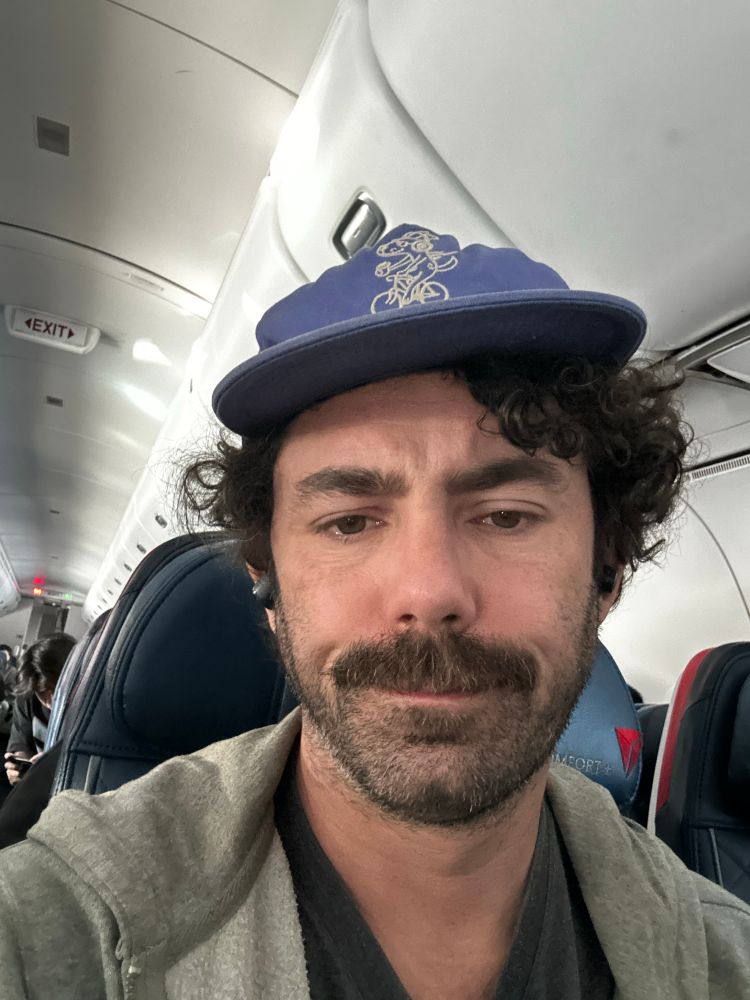 Selfie of a grizzled yet youthful 42yo man on an airplane. He has a tight, disdainful look on his face despite having consumed two martinis before boarding 