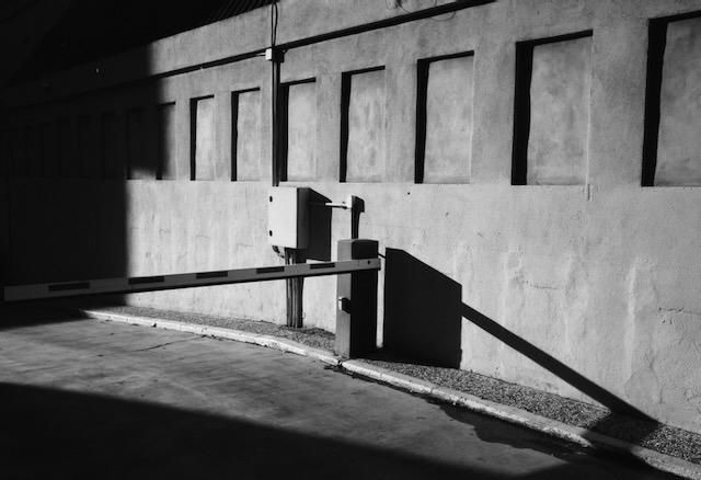 Una foto en blanco y negro donde se ve una pared con vanos cerrados que generan un ritmo visual armónico. Una valla de elevación automática, cerrada para el paso de vehículos, un alto contraste entre luz y sombra.