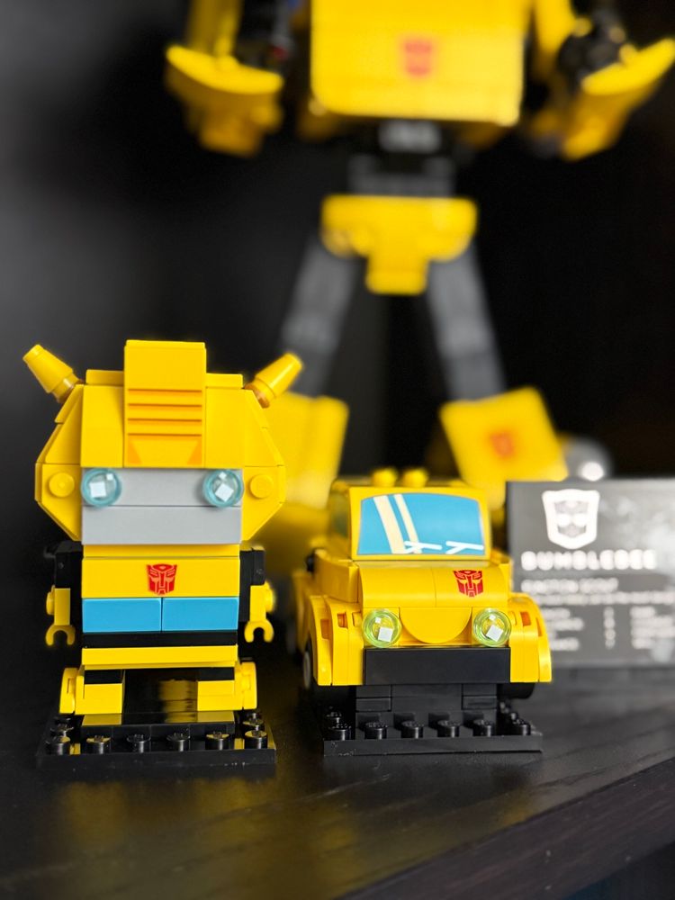 Lego Transformer Bumblebee robot and car Brickheadz set standing in front of a larger Lego Icons Bumblebee Lego build on a bookshelf next to a bumblebee placard. 