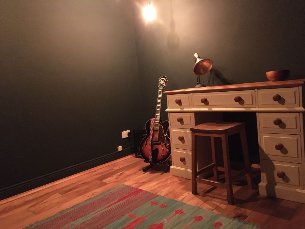 A newly decorated room with a desk and stool, an electric guitar, a stripped wooden floor and a blue / green and red rug. The walls are very dark green.