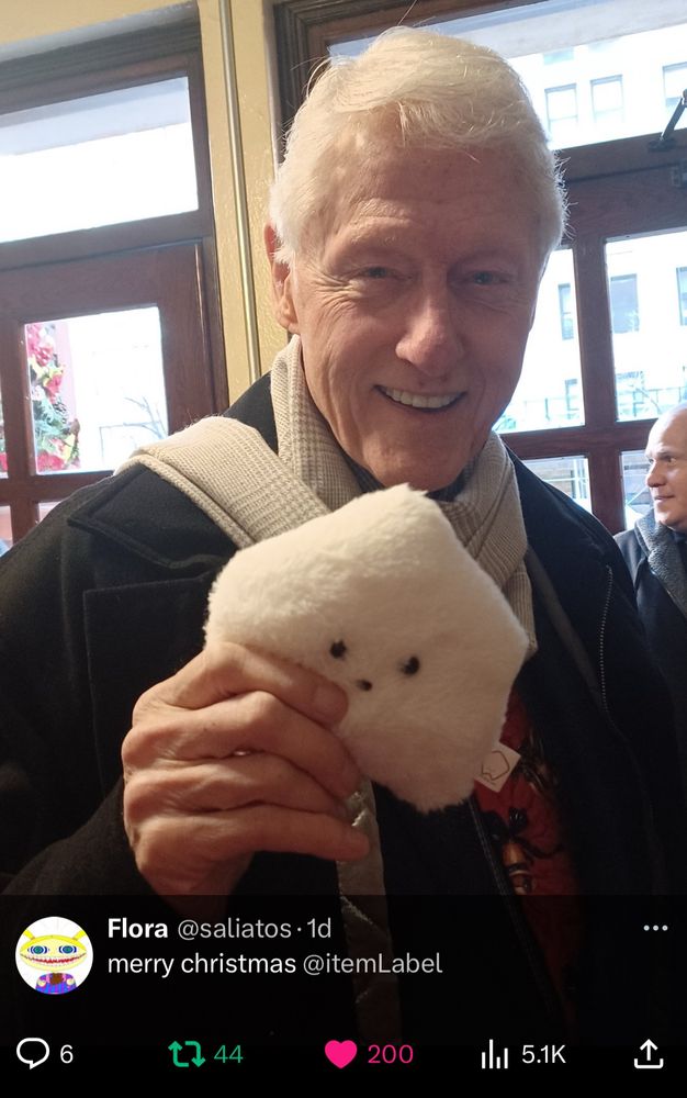 Former Presidential Bill Clinton smiling and holding a Sucklet plush towards the camera, screenshot from a recent tweet by @/saliatos