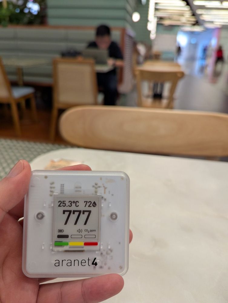 Me sitting down in a dining area of a mall, holding a CO2 Sensor (Aranet Home 4)
