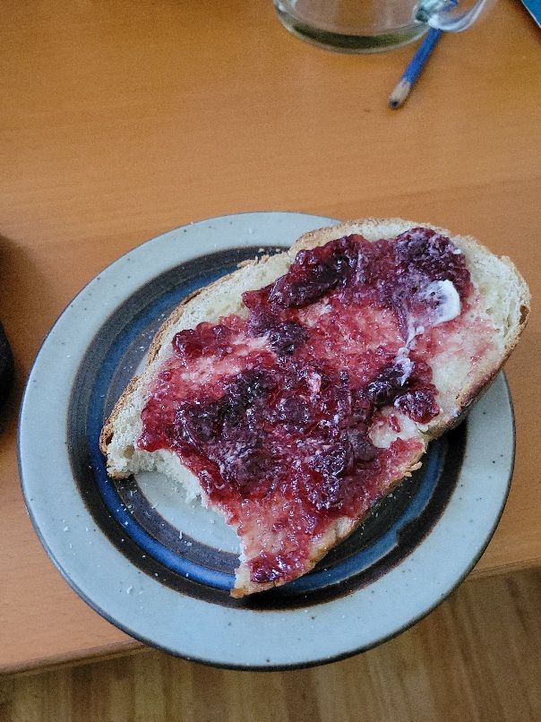 A rounded slice of light brown rye bread with red cherry jam on it. It's on a beige, brown, & blue plate.