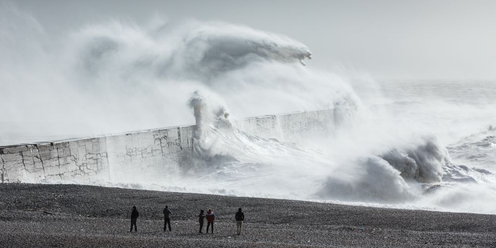 A photograph of a monstrous storm wave crashing over a sea wall watched by 5 tiny figures on the beach. Newhaven, East Sussex, UK
