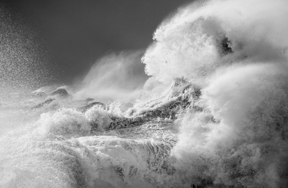 A black and white photo of a breaking wave (on right) and spray in foreground (on left). 