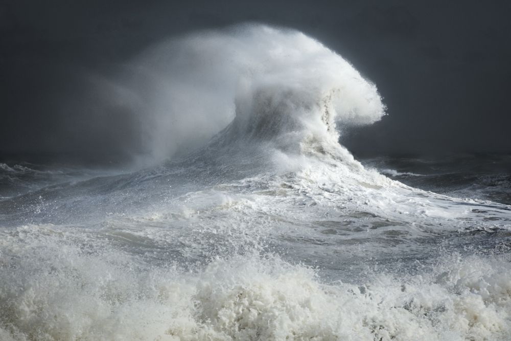 Two clashing storm waves form a strange shape and white spray against a dark blue sky. 
