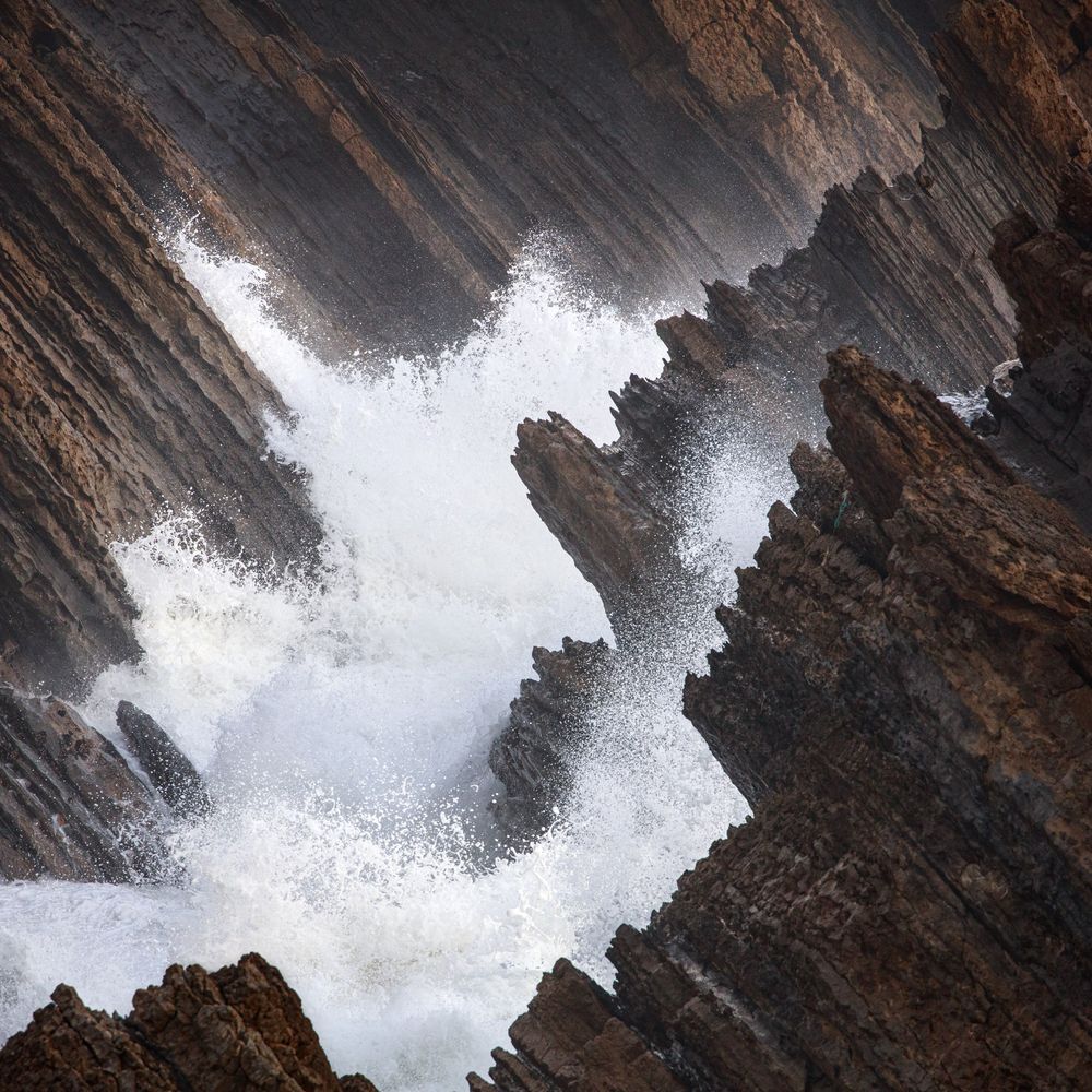 A square photo of jagged brown rocks and foamy sea water.