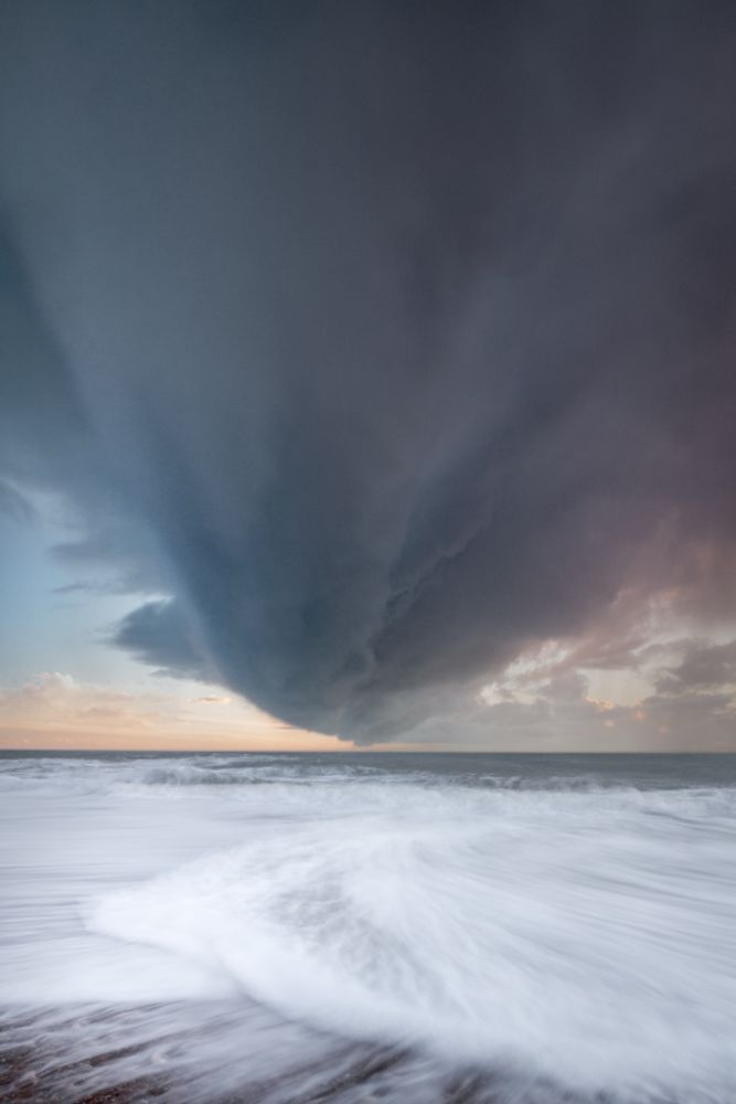 A vertical-format photo of a funnel-shaped cloud. The horizon is below centre. In the foreground, some foamy sea waves cross each other on the shore. 