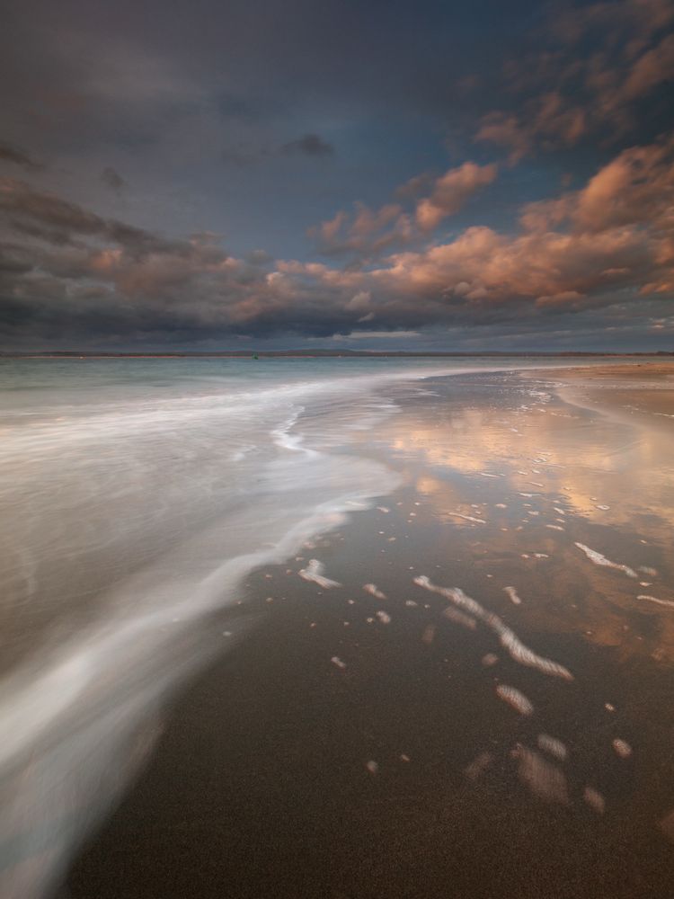 A vertical format photo of a sandy beach at sunset. Horizon above centre. A line of blurred sea foam leads the eye from bottom left. Pink colours are reflected in the wet sand. 