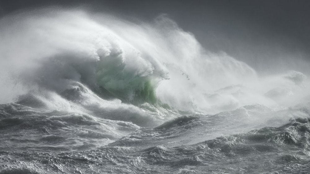 A photo of a curling storm wave on left of frame, mostly grey with a flash of backlit green. Copious spindrift against a dark, brooding sky. 