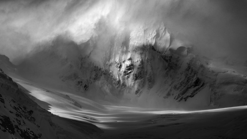 A black and white photo of a mountain in Antarctica. Shadowy foreground gives way to the mountainside, much spindrift blowing off the face of the mountain, a beam of light from the left. 