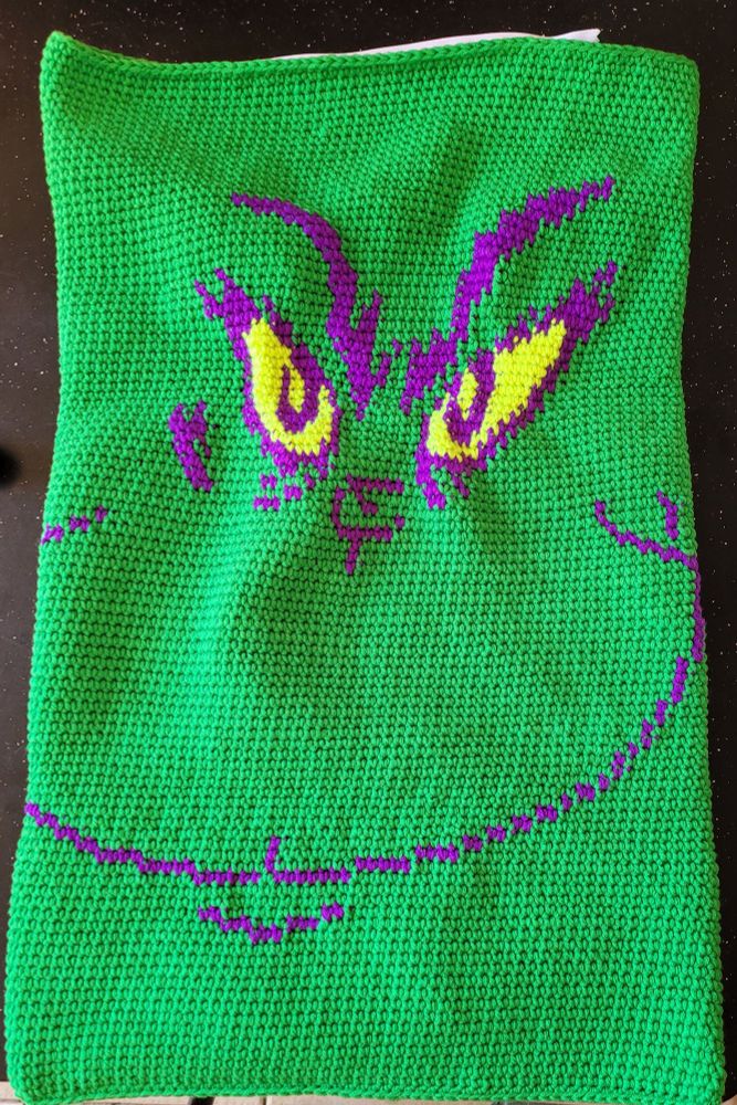 Crocheted pillow case. Main colour is leaf green. Purple is used for the mouth, nose, eyebrows, fur tufts around the eyes, and outlining the eyes. The eyes are fluro yellow. 