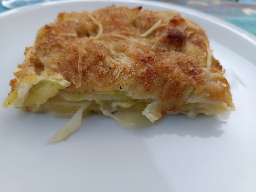 A piece of pie on a plate. The surface is browny the content is green and white. It's a pie made of cheese, potatoes and trombetta squash lies. 