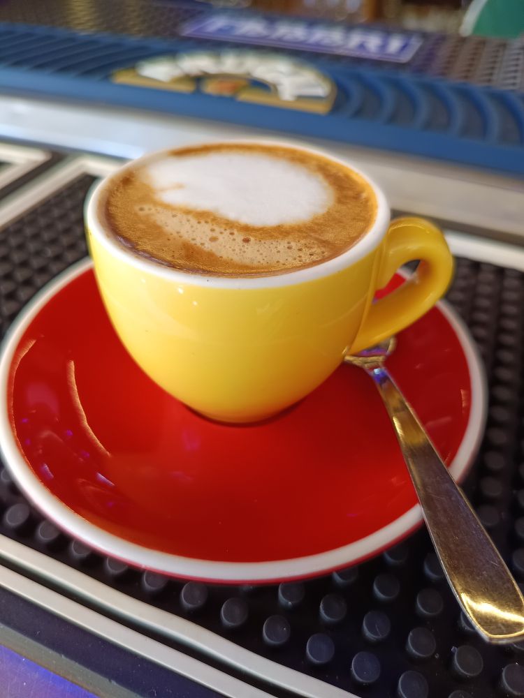 A cup of espresso macchiato. The cup is yellow, the small plate red, a metal spoon.