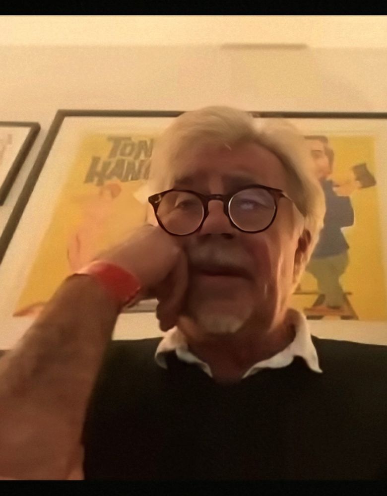 A man in his late 60s wearing heavy framed glasses. He is in close up and appears relaxed with his right hand raised to his cheek, lightly resting his face against the back of his finger. He has a grey moustache, a small goatee beard and a full head of grey hair. He is talking. The man is Glen Matlock.