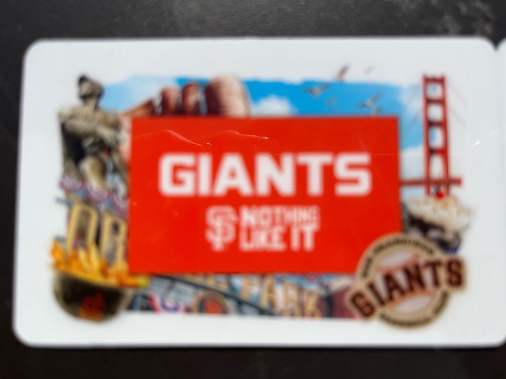 A white library illustrated with a collage of the Golden Gate Bridge, the Willie Mays statue, a baseball helmet filled with garlic French fries, and a San Francisco Giants baseball club logo. White text on an orange banner in the center reads “Giants, Nothing Like It”