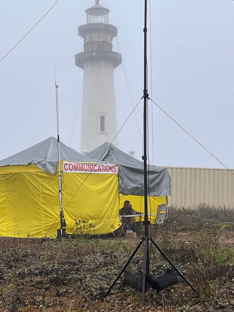 A bright yellow tent with a banner reading “Communications” sits in a field in front of a tall shite historic lighthouse. In the foreground is a tall black vertical radio antenna. The sky is foggy gray.