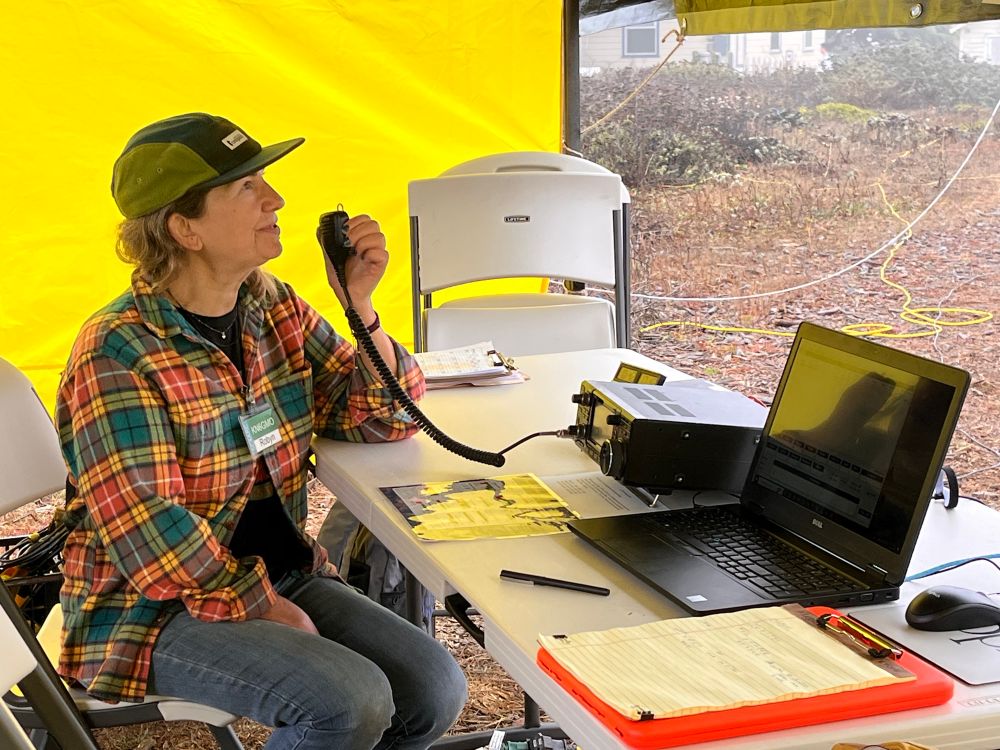 A woman in a brightly colored plaid shirt and ball cap sits at a table in front of an amateur radio set and a laptop. She has the radio microphone in her hand and is smiling. 
