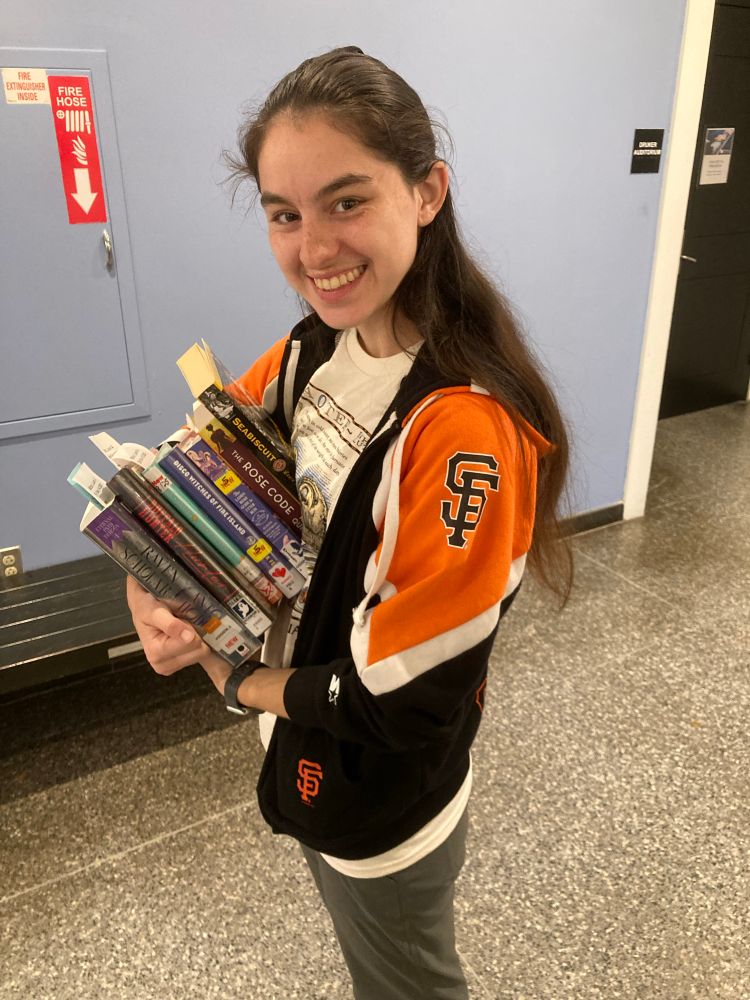 A young woman in a black and orange jacket stands sideways to the camera. She has an armload of library books and she is smiling. 