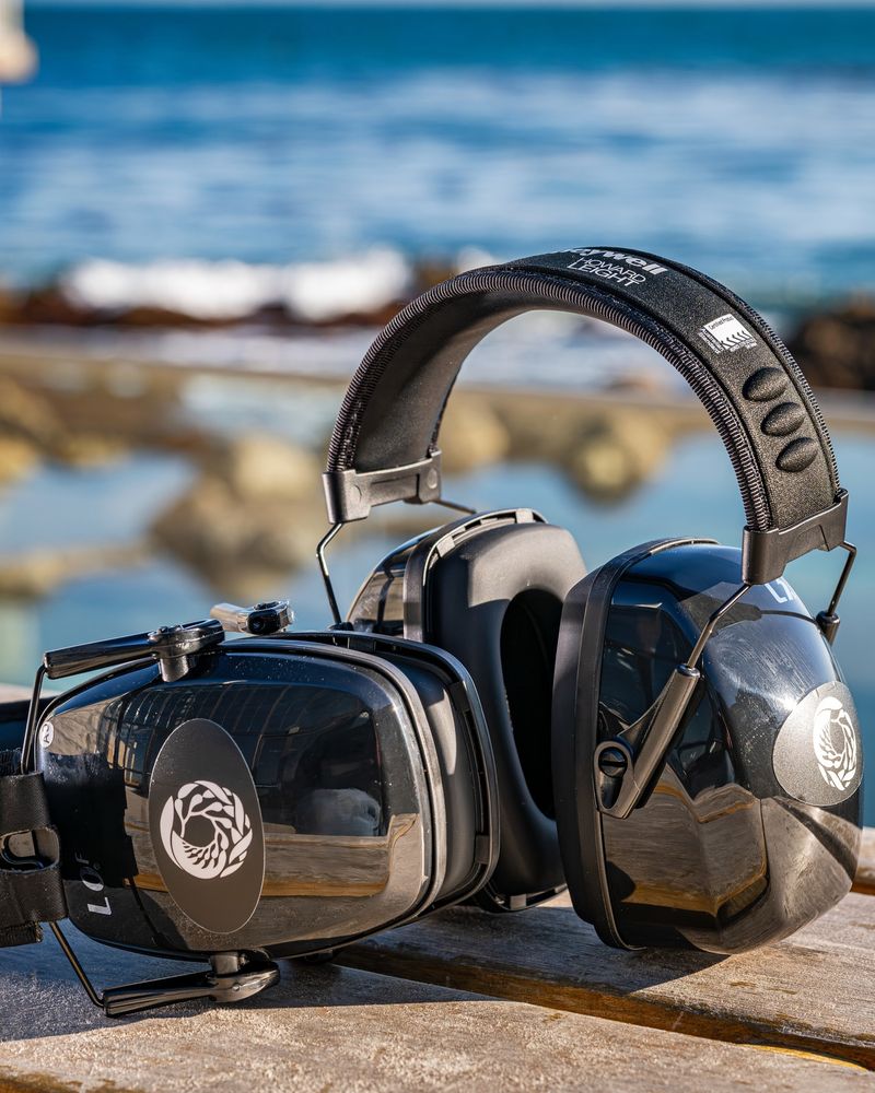 A pair of over-ear sensory headphones with a white Monterey Bay Aquarium logo rests on a wood surface with the ocean in the background