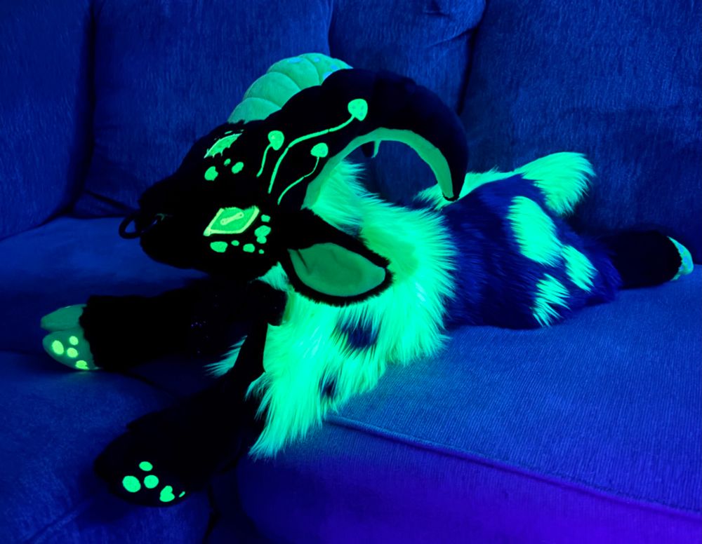 Black and green goat plush laying on a couch. Green is UV blacklight reactive and designed to look like fungi is overtaking the goat. 