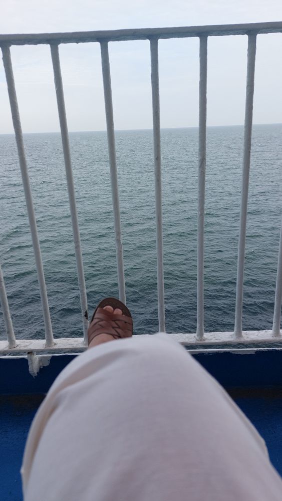 Gitter mit Blick auf Meer. Im unteren Rand ein Weiße Hose und ein Fuß in brauner Sandale