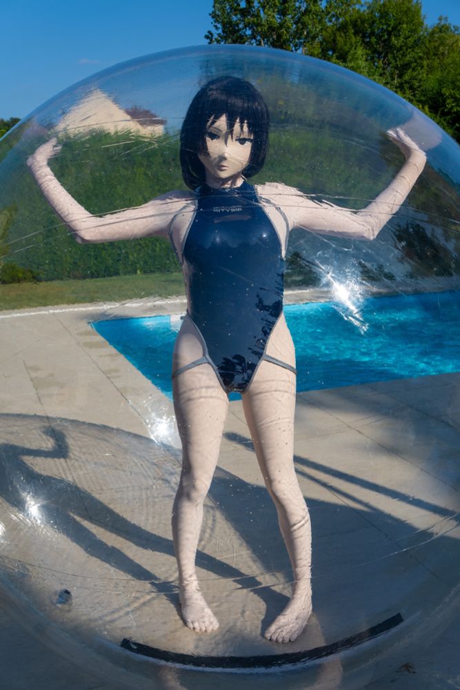 Oboro, wearing a navy blue Bitysie swimsuit, is standing in a big inflatable transparent ball on the edge of a pool. The sunlight is reflecting off the ball.