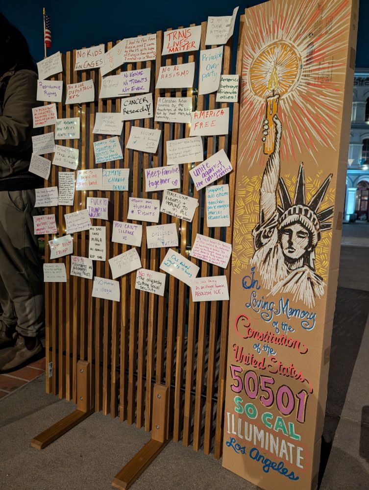 Outdoor photo at night shows a wooden slatted board covered in small white papers with handwritten messages, alongside a large cardboard sign with a drawing of the Statue of Liberty holding a lit candle. The sign reads "In Loving Memory of the Constitution of the United States" Various messages on the papers include "TRANS LIVES MATTER," "No Kids in Cages," "No Al Rasismo," and others expressing social and political sentiments.