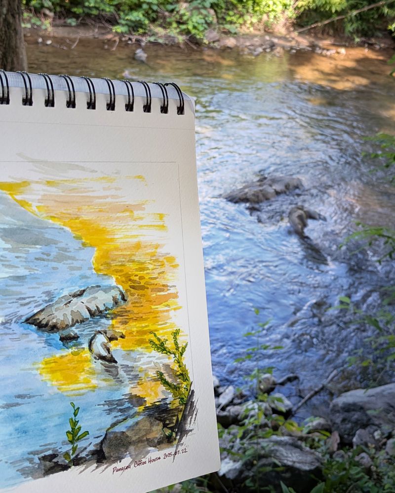 Photo of the watercolor painting and actual subject in view: rocks in a stream with golden sunlight casting a diagonal cool/warm partition across the image.