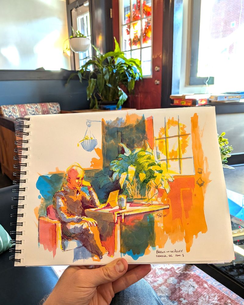 Portrait of a man in a coffee shop sketched in high chroma ink. A table attached to a wooden chair and a potted plant block a red door, but in the sketch it is a bright orange. A hanging plant is in the background. The man is watching a tablet, but has left by the time the drawing is completed.