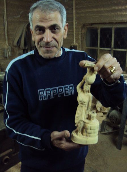 a middle eastern man holding a highly detailed figurine of a woman carrying a jug on her head standing next to a well.  He's wearing a track suit top that inexplicably has the word RAPPER printed on it