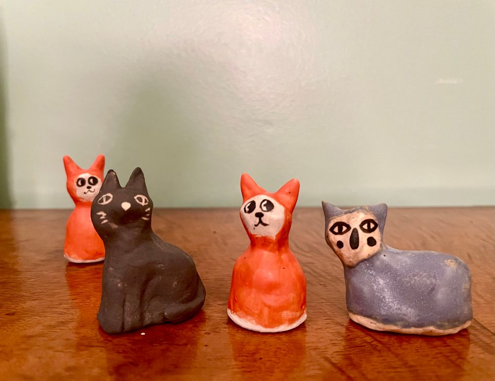4 small ceramic figures sit on a orange brown wood veneer surface with a light blue-green wall background. 2 orange cat figures, 1 black cat figure & one blue sort of cat-like figure. 
