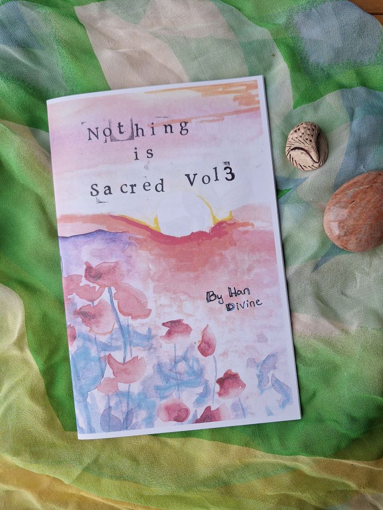 A zine against a green and yellow silk scarf. An owl worry stone and peach moonstone are also on the cloth. The zine is a brilliantly bright yellow and pink sunset over an orange hill with red and blue abstract poppy flowers  in the foreground. In typewriter set it reads 'Nothing is Sacred vol 3 By Han Divine'.