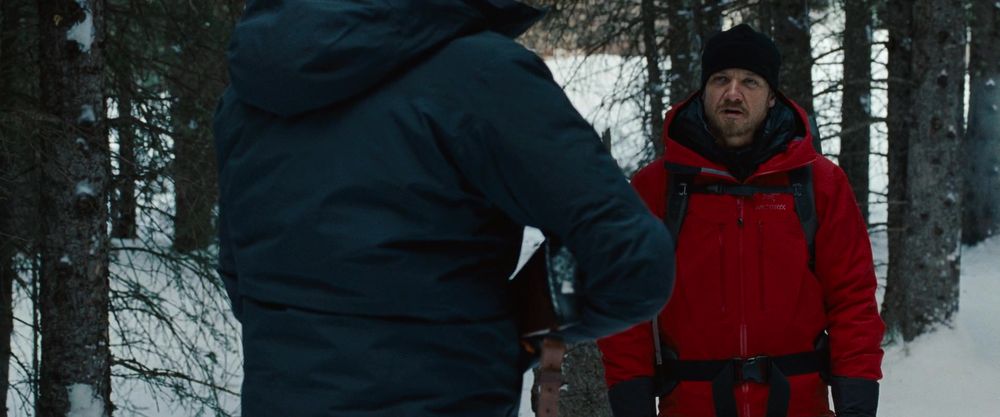 Aaron Cross, portrayed by Jeremy Renner, speaking with Outcome #3. He is wearing a red parka and is not holding a gun. 