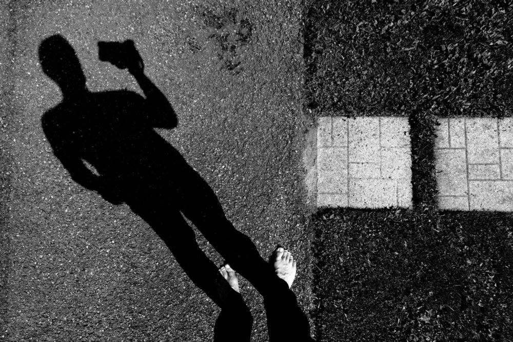 A stark black-and-white photograph taken from above, where shadow, texture, and human presence intertwine. At the center-left of the frame, the elongated silhouette of a person stretches across rough asphalt, cast sharply by a high sun. The figure is holding a camera or phone in their hand, the device raised, capturing the very scene we are now observing, a reflexive moment where subject and photographer collapse into one. Below, just outside the shadow’s reach, bare feet stand firmly on the ground, toes splayed naturally, grounding the image in both vulnerability and authenticity. To the right, the composition shifts: two stone tiles break through the dark patch of grass, squares of order interrupting the organic chaos of nature. The play of light and darkness defines the atmosphere: the shadow is deep and heavy, almost ink-black, while the textures of asphalt, grass, and stone emerge in crisp detail. Emotionally, the image speaks to self-awareness and presence, the artist revealed not by face but by absence, by shadow. It is a quiet meditation on perception, existence, and the intimate connection between photographer, subject, and the simple act of standing still to observe. The high-contrast tones enhance the surreal quality, turning the ordinary into a statement on perspective. This “shoot from above” is not only about the view downward but also about looking inward, confronting the delicate intersection between identity and the world beneath one’s feet.
