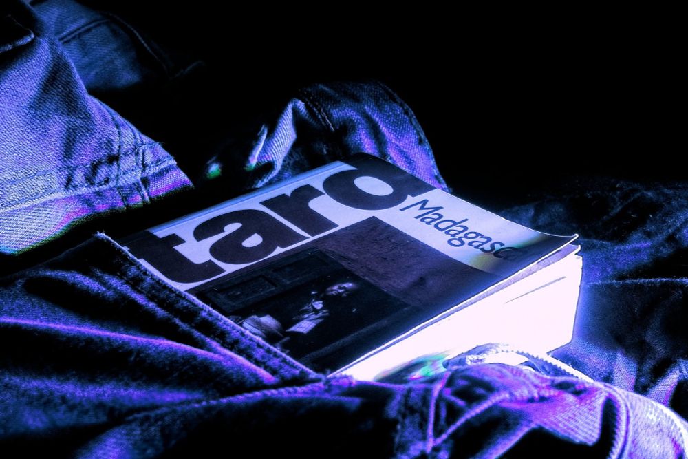 In the photograph, a French travel guidebook, Le Guide du Routard - Madagascar, lies partly tucked into a dark denim pocket, its thick pages catching a cold blue-violet light. The bold word “routard” dominates the cover like a headline to adventure, paired with “Madagascar,” a name that conjures distant islands, winding roads, and red-earth horizons. The textured jeans, with their seams and folds, evoke rugged terrain and dusty tracks, as though the fabric itself were a map of yet-to-travel paths. The ultraviolet glow gives the scene an otherworldly feel, like neon light spilling from a roadside motel at dusk or the glow of a dashboard during a night journey. The cover photo of a person in a doorway, printed small beneath the title, brings a human presence and a hint of intimacy to the otherwise bold, graphic cover, an image of the everyday life one might encounter on the road. This combination of textures, typography, and lighting suggests the liminal space between preparation and departure: the quiet moment before the road trip begins, when maps are folded and guides are slipped into pockets. The photograph speaks to the theme “The Road” not only as a literal path across Madagascar but as a state of mind: freedom, curiosity, anticipation, and the soft hum of possibility. The angle of the shot, looking down at the guidebook as though seated in transit, echoes the feeling of holding your own belongings on a train, a bus, or at a remote roadside café, poised on the edge of movement. It’s a portrait of wanderlust distilled into a single object: a guidebook glowing like a talisman under electric light, promising encounters, detours, and memories to come.