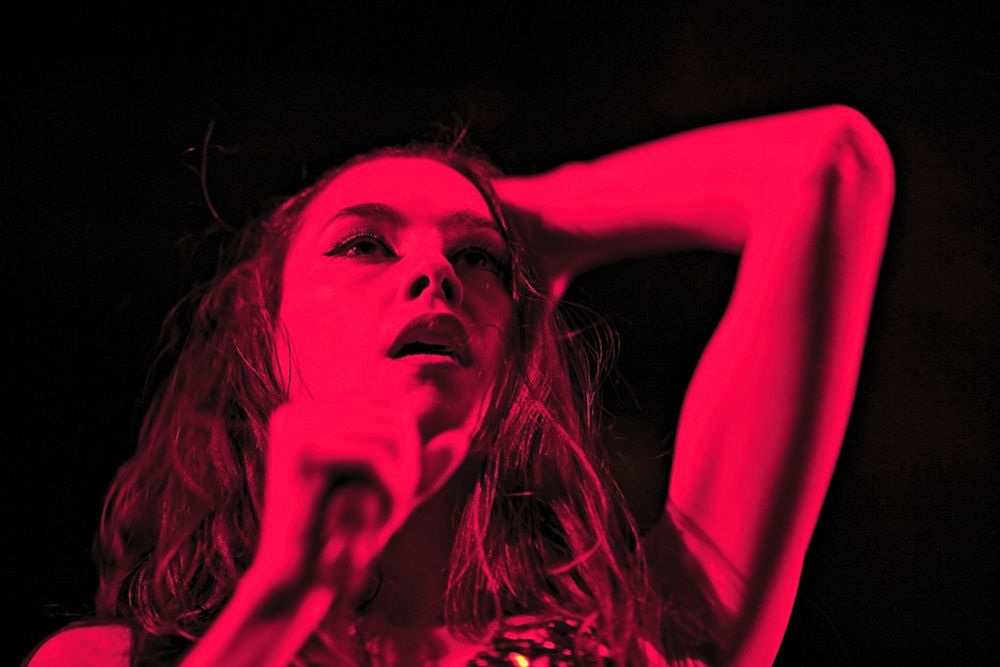 A striking close-up of a female performer mid-song, bathed in an intense, saturated magenta-red stage light that floods the entire frame with electric energy. Her expression is raw and evocative—lips parted slightly as if caught mid-note, eyes lifted toward the stage lights or perhaps something more abstract, capturing the ephemeral moment where emotion and performance blur. One arm is raised behind her head, a gesture that enhances both the drama and sensuality of the image, while the other grips a microphone in the foreground, slightly out of focus, adding to the immediacy and immersion of the viewer’s perspective.

Her hair, tousled and damp with sweat, glistens subtly under the colored lighting, emphasizing the physical intensity of the performance. The background is a deep, enveloping black, which makes the vibrant red tones even more explosive and visually dominant—there is no visual distraction, only her presence, luminous and commanding. Highlights carve sculptural lines along her face and arm, further dramatized by the chiaroscuro effect of the lighting.

The atmosphere pulses with life and movement, as if the very air around her is humming with the rhythm of the music. This photograph doesn’t just suggest vibrancy—it radiates it. It’s a visual symphony of passion, color, and sonic intensity frozen in a single frame, celebrating the fierce vitality of live performance and the magnetic power of a performer caught in a moment of unfiltered artistic release.