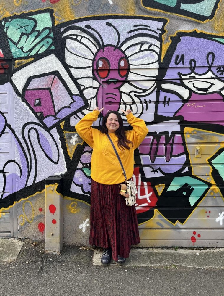 A girl in a yellow jumper standing under graffiti of the Pokémon butterfree
